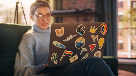 Stylish Female Using Laptop Computer with Diverse LGBT and Lifestyle Stickers on the Back. Young Creative Woman Sitting on a Couch, Browsing Internet, Checking Social Mediaの写真素材