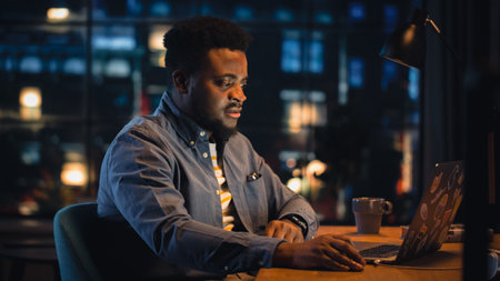 Young Handsome Black Man Working from Home on Laptop Computer in Stylish Loft Apartment in the Evening. Creative Male Gets a Great Idea while Checking Social Media. Urban City Viewの写真素材