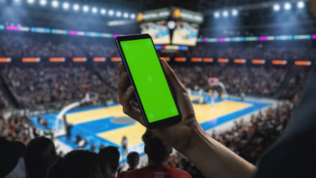 Basketball Championship: Person's Hand Holding a Smartphone with a Green Screen Display. Sports Fans in an Arena Cheering for a Team to Win. Template to Use for Social Media,の写真素材