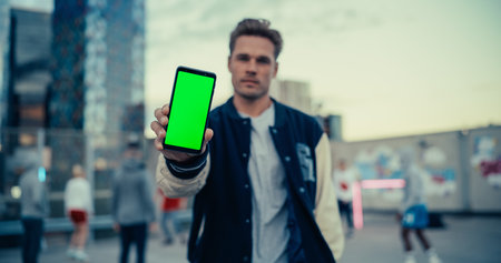 Handsome Caucasian Man Holding a Smartphone, Showing the Green Screen Mock Up Display. Young Stylish Man Standing in a City Environment, Diverse People Playing Street Soccerの写真素材