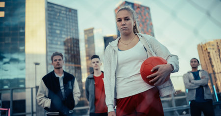 Empowering Portrait of a Beautiful Sporty Young Woman Looking at Camera, Holding a Red Soccer Ball in Her Hands. Stylish Female Footballer Standing Behind a Fence with a Groupの写真素材