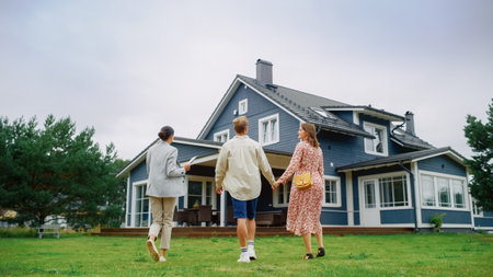 Real Estate Agent Showing a Beautiful Big House to a Young Successful Couple. People Standing Outside on a Warm Day on a Lawn, Talking with Businesswoman, Discussing Buyingの写真素材