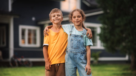 Young Happy Brother and Sister Standing in Front of a Country House, Embracing Each Other, Looking at Camera and Smiling. Two Kids Enjoyingの写真素材