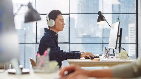 Young Asian Man Working on a Computer and Listening Through Headphones in a Modern Office. Male...