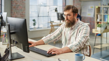 Portrait of Young Caucasian Bearded Man with Glasses Focused While Sitting and Working on Computer...