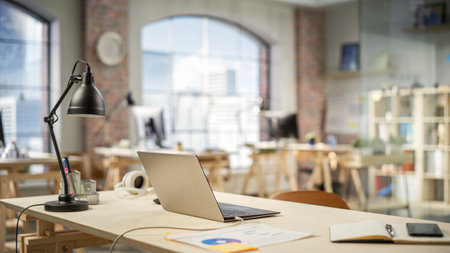 Charging Laptop on a Working Station in a Bright Spacious Empty Workplace. Office Supplies Are on the Table. Big Windows with City Buildings Viewの写真素材