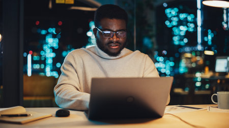 Stylish Black Specialist Sitting at a Desk in Creative Agency in the Evening. Young Handsome African...