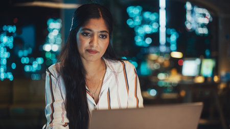 Close Up Portrait of a Happy Indian Manager Sitting at a Desk in Creative Office. Young Stylish Female with Long Dark Hair Using Laptop Computer in Marketing Agencyの写真素材