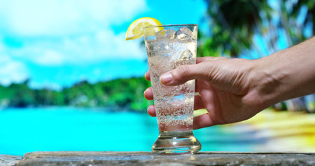 man hand holding a glass of water with ice and lemon on the beachの写真素材