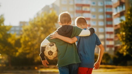 Two Young Caucasian Boys Embracing Each Other while Walking to a Neighborhood Football Pitch in Sport Clothes. Soccer Players Carrying a Ball to Play. Concept of Sports, Childhood, Friendship.の写真素材