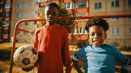 Portrait of Two Young Talented African Boys Standing Together while Looking at Camera and Smiling. Older Brother Holding a Soccer Ball. Black Kids Standing in Front of a Football Gate.の写真素材