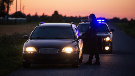 Highway Traffic Patrol Car Pulls over Vehicle on the Road. Female Police Officer Approaches Driver Asks for License and Registration. Black Officer of the Law doing Job Professionally. Side Back Viewの写真素材