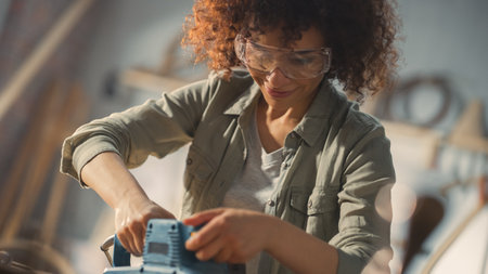 Female Carpenter Wearing Protective Safety Glasses and Using Electric Belt Sander to Work on a Wood...