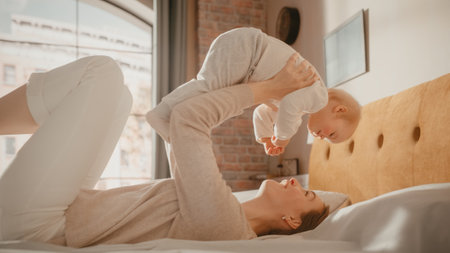 Caring Mother Lying on a Bed, Playing with a Cute Newborn Baby at Home. Mom Bonding with a Toddler, Holding Happy Child Above Her. Concept of Childhood, New Life, Motherhood.の写真素材