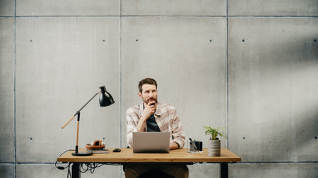 Professional Creative Man Sitting at His Desk in Office Working on a Laptop Computer, Working, Thinking, Solving Problems and Smiling at Success. Stylish Office Space Design Perfect for Productivityの写真素材