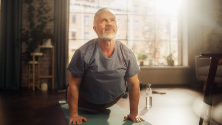 Portrait of a Strong Fit Senior Man Training on a Yoga Mat, Doing Back Stretching and Core Strengthening Exercises During Morning Workout at Home in Sunny Apartment. Concept of Health and Fitness.の写真素材