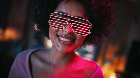 Close Up Portrait of Multiethnic Young Black Latin Female with Afro Hair in Futuristic Neon Glowing Glasses, Having a Fun at Home, Smiling and Posing for Camera in Loft Apartment in the Evening.の写真素材