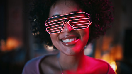 Close Up Portrait of Multiethnic Young Black Latin Female with Afro Hair in Futuristic Neon Glowing Glasses, Having a Fun at Home, Smiling and Posing for Camera in Loft Apartment in the Evening.の写真素材