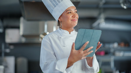Restaurant Kitchen: Black Female Chef Cooking Delicious and Authentic Food, Uses Digital Tablet Computer. Professional Creates traditional Dish, using Healthy Ingredients for Recipe Preparationの写真素材
