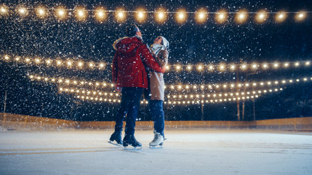 Romantic Winter Snowy Evening: Ice Skating Couple Having Fun on Ice Rink. Pair Figure Skating at Beautifully Lit Location. Boyfrined Meeting Girlfriend, Starts Dancing. Tender Young People in Loveの写真素材
