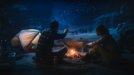 Happy Couple of Hikers Camping in the Canyon at Night Together, Sitting by Campfire, Plan Hiking Trekking Journey Route with a Map. Two Traveling people Talk, Milky Way Star Watching. Vacation Tripの写真素材