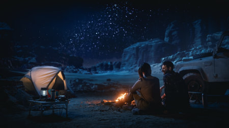 Happy Couple Tent Camping in the Canyon, Sitting by Campfire Watching Night Sky with Milky Way Full of Bright Stars. Two Travelers In Love On a Romantic Vacation Trip. Back View Shotの写真素材