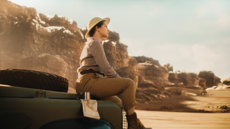 Desert Road Trip: Beautiful Female Traveler Looking Around while Sitting on a Hood of Her Offroad SUV, Planning Route full of Wonder and Exploration. Woman Adventurer Traveling Through Canyonの写真素材