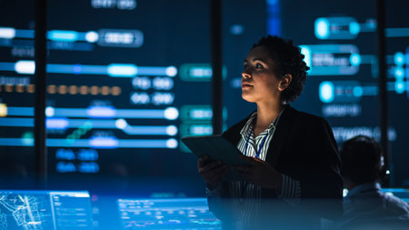 Young Multiethnic Female Government Employee Uses Tablet Computer in System Control Monitoring Center. In the Background Her Coworkers at Their Workspaces with Many Displays Showing Technical Data.の写真素材