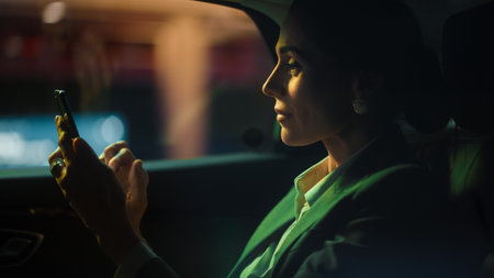 Beautiful Businesswoman is Commuting from Office in a Backseat of Her Luxury Car at Night. Entrepreneur Using Smartphone while in Transfer Taxi in Urban City Street with Working Neon Signs.の写真素材