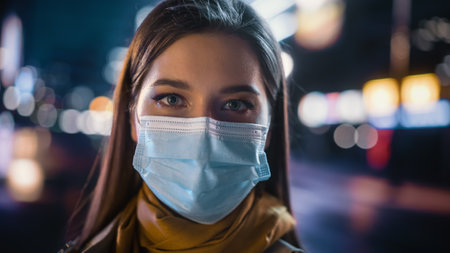Portrait of a Beautiful Woman Wearing a Protective Face Mask in Trench Coat Standing in a Modern City Street with Neon Lights at Night. Female Looking at Camera in the Urban Cinematic Environment.の写真素材