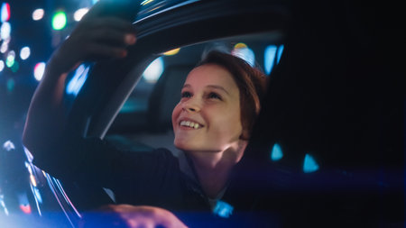 Excited Young Boy is Looking Out of the Window from Backseat of a Car at Night. Kid Taking Photos and Videos on a Smartphone with Amazement of How Beautiful is the City Street with Working Neon Signs.の写真素材