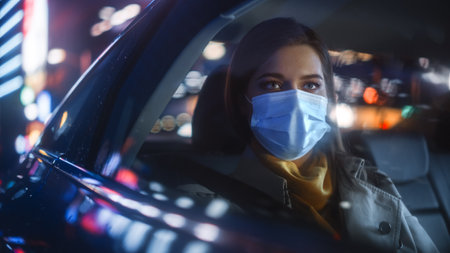 Female Wearing Face Mask is Commuting Home in Backseat of a Taxi at Night. Beautiful Female...