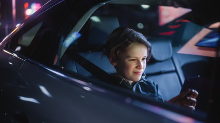 Young Boy is Sitting on Backseat of a Car, Commuting Home at Night. Passenger Watching Funny Entertainment on Smartphone while in Taxi in City Street with Working Neon Signs.の写真素材