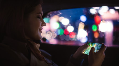 Happy Female is Commuting Home in a Backseat of a Taxi at Night. Beautiful Woman Passenger Playing Strategy Video Game on Smartphone while in a Car in Urban City Street with Working Neon Signs.の写真素材