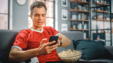 Charismatic Young Adult Man Sitting on a Couch Watches Game on TV, uses Smartphone App for Score, Bet, Statistics, Celebrates Victory when Team Wins Championship. Happy Fan Watches Sport.の写真素材