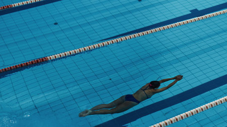 Beautiful Female Swimmer Diving in Swimming Pool. Professional Athlete Jumped From Starting Block, Swims under Water. Person Determined to Win Championship. High Angle View.の写真素材