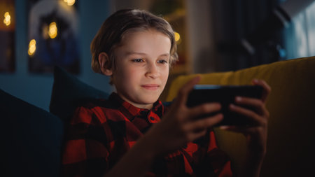 Cute Young Boy Sitting on a Sofa in a Dark Cozy Room and Using a Smartphone at Home. Happy Teenager...