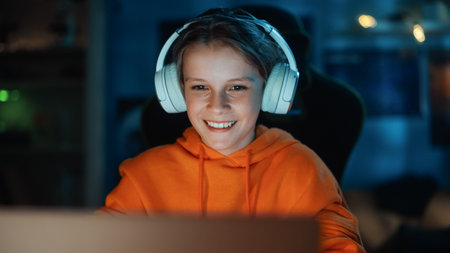 Smart Young Boy in Headphones Using Laptop Computer in Cozy Dark Room at Home. Happy Teenager Browsing Educational Research Online, Chatting with Friends, Studying School Homework.の写真素材