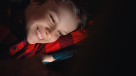 Cute Young Boy Lying in Bed in a Dark Room and Using a Smartphone at Home. Happy Teenager Browsing...