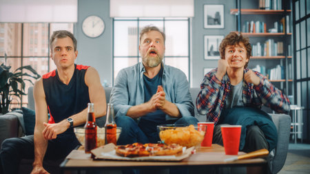 At Home Three Joyful Basketball Fans Sitting on a Couch Watch Game on TV, Celebrate Victory when Sports Team Wins Championship. Group of Friends Cheer When Favourites Play.の写真素材