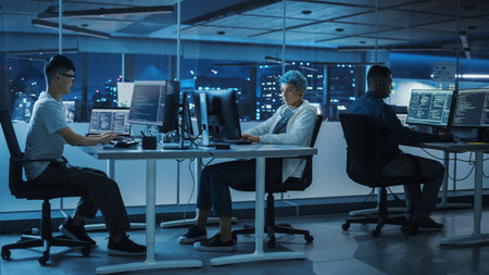 Diverse Office at Night: Multi-Ethnic Team of Young Programmers Working on Desktop Computers,...