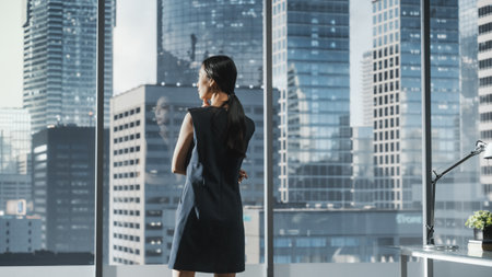 Successful Businesswoman in Stylish Dress Looking out of the Window at Big City in Downtown Area. Confident Female CEO Working on Financial Projects. Manager at Work Planning Marketing Campaign.の写真素材