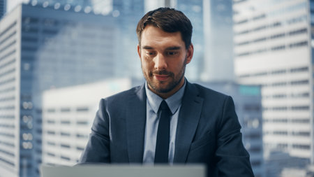 Smiling Businessman in a Suit Sitting at a Desk in Modern Office, Using Laptop Computer, Next to...
