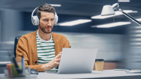 Modern Office: Handsome Businessman Sitting at His Desk Working on a Laptop Computer. Man Wearing Headphones, Listens to Music, Podcast. Motion Blur Background Showing Active Work Day.の写真素材