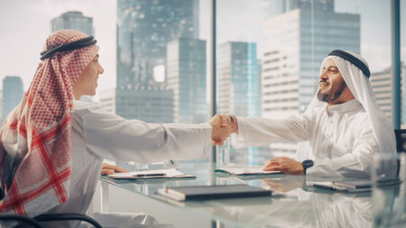 Two Joyful Emirati Businessmen in White Traditional Kandura Sitting in Office and Signing Contract....