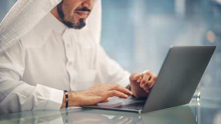 Successful Arab Businessman in White Traditional Outfit Sitting in Office and Working on Laptop Computer. Business Manager Planning Corporate Strategy. Saudi, Emirati, Arab Businessman Concept.の写真素材