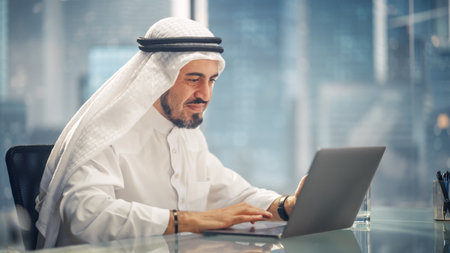 Arab Businessman in White Traditional Outfit Sitting in Office and Working on Laptop Computer. Business Manager Make Successful Investment Deal. Saudi, Emirati, Arab Businessman Concept.の写真素材