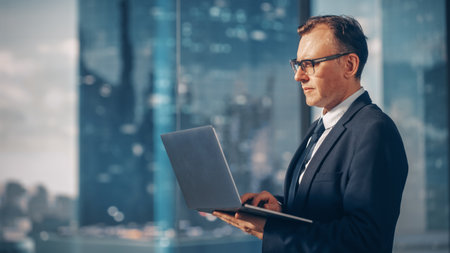 Confident Businessman in a Suit Standing in Modern Office, Using Laptop Computer, Looking out of the...