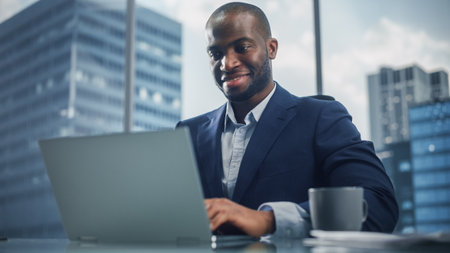 Portrait of Successful Black Businessman in Tailored Suit Working on Laptop Computer in His Big City Office. Digital Entrepreneur does Data Analysis, Cloud Computing for e-Commerce Strategy assessmentの写真素材