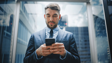 Successful Businessman Riding Glass Elevator to Office in Modern Business Center. Handsome Happy Man Smile while Using Smartphone, Write Text Messageの写真素材